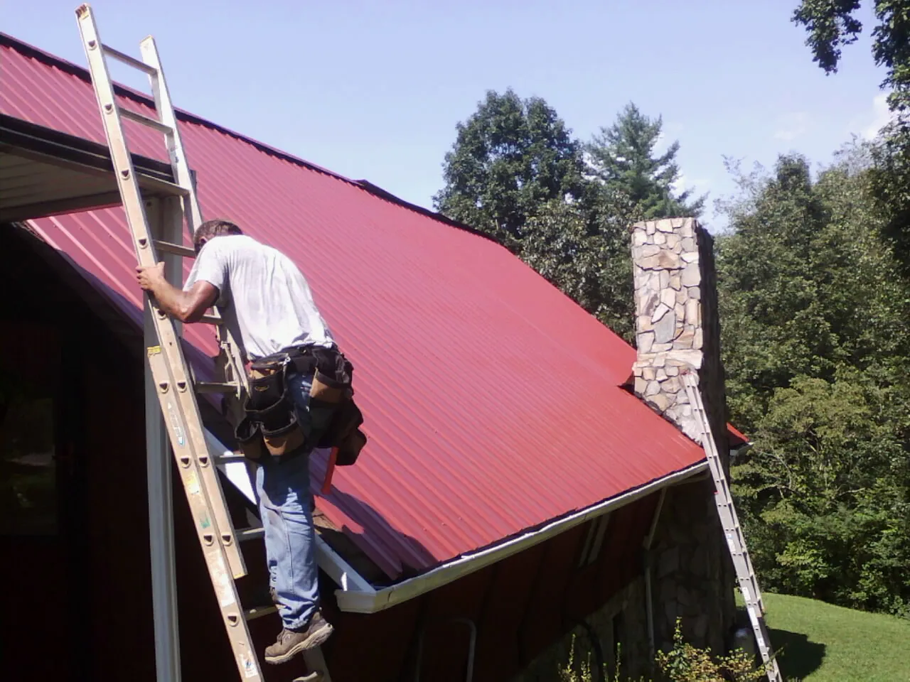 Roofer on a ladder beside a red metal roof and stone chimney — Soffit & Fascia Repair by Roofing Brothers in Mount Crawford